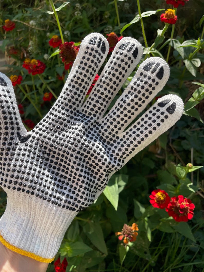 Op de afbeelding: Een witte gebreide handschoen met een zwart stippenpatroon voor grip op de handpalm en vingers, met zwarte pootafdrukken op de toppen van elke vinger. De handschoen heeft een gele geribbelde boord bij de pols voor een goede pasvorm.