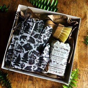 Op de afbeelding: Een geschenkdoos met een witte schoonmaakhandschoen met een zwart patroon van padden, paddenstoelen en varens, een houten schrobborstel met lichte haren en een stuk Toad-Ally Clean rozemarijn-muntzeep met een label waarop 114g staat.