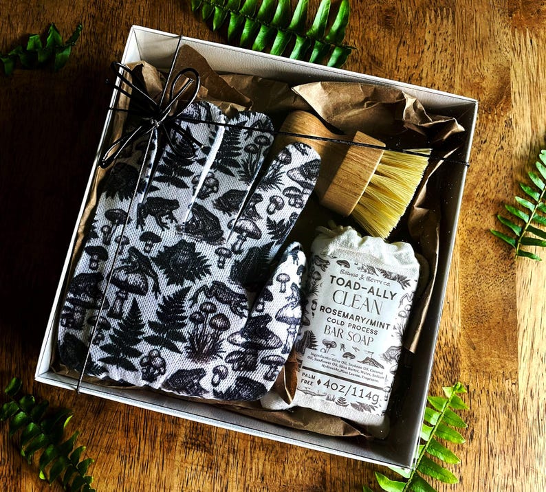 Op de afbeelding: Een geschenkdoos met een witte schoonmaakhandschoen met een zwart patroon van padden, paddenstoelen en varens, een houten schrobborstel met lichte haren en een stuk Toad-Ally Clean rozemarijn-muntzeep met een label waarop 114g staat.