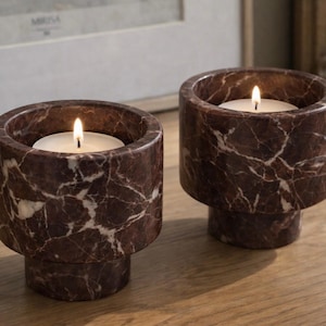 Pode incluir: Dois castiçais de mármore com velas acesas. Os castiçais são cilíndricos com uma base de pedestal, apresentando um padrão marmorizado marrom e branco. As velas são brancas e as chamas são brilhantes.