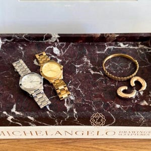 May include: A rectangular marble tray with a burgundy and white pattern holds a silver watch, a gold watch, a gold bracelet, and gold earrings. The tray has the text "MICHELANGELO DRAWINGS SCULPTURES" on the front.