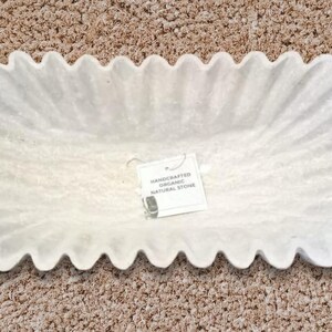May include: White oval-shaped serving dish made of natural stone with a scalloped edge. The dish has a handmade organic look. The label reads "Handcrafted Organic Natural Stone".