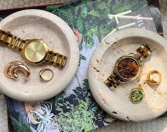 Puede incluir: Dos bandejas de piedra beige sostienen cada una un reloj dorado, joyas y accesorios. Una bandeja tiene un reloj dorado, un par de pendientes de aro dorados y un anillo dorado. La otra bandeja tiene un reloj dorado, un anillo dorado y un colgante verde.