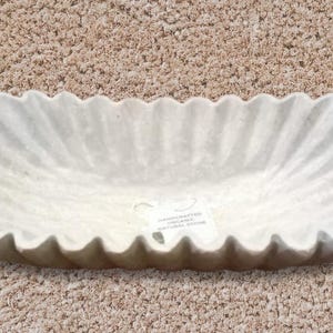 May include: A white oval-shaped ceramic planter with a fluted edge. The planter has a textured surface and a label on the bottom.