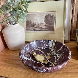 May include: A red and white marble bowl with a gold watch and chain inside. The bowl is sitting on a wooden surface.