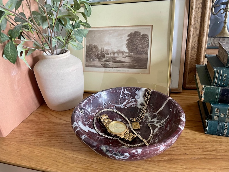 May include: A red and white marble bowl with a gold watch and chain inside. The bowl is sitting on a wooden surface.