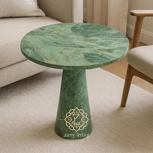 Könnte beinhalten: Ein runder, grüner Beistelltisch aus Marmor mit einem einzelnen, konischen Fuß. Die Tischplatte hat ein wirbelndes Muster aus Grün und Weiß. Der Sockel hat ein goldenes Emblem und den Text "zany living". Der Tisch hat einen Durchmesser von ca. 50 cm.
