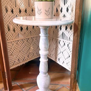 May include: A white marble-look pedestal table with a round top. The table supports a small potted succulent plant. The table has a decorative, turned column base and a circular foot. The background includes a macrame screen and a teal wall.