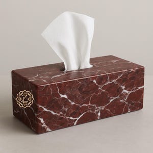 Pode incluir: Uma caixa de lenços retangular feita de mármore vermelho com veios brancos. Um emblema dourado com a letra "L" está na frente. Lenços brancos saem da abertura superior. Esta caixa decorativa adiciona um toque de elegância a qualquer ambiente.
