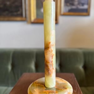 May include: A paper towel holder crafted from green and brown onyx. It features a circular base, a cylindrical central column, and a rounded top. The onyx displays natural veining and patterns. The holder is placed on a wooden surface.