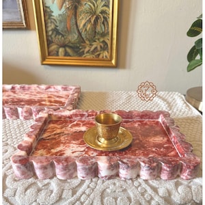 Peut inclure: Deux plateaux rectangulaires avec des bords festonnés et un motif de marbre rouge et blanc. Une petite tasse et une soucoupe dorées sont posées sur un plateau. Les plateaux sont sur une nappe en dentelle blanche. Une peinture est accrochée en arrière-plan.