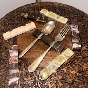 Puede incluir: Una colección de seis reposa palillos rectangulares de piedra y madera con ranuras, que presentan patrones variados en tonos verdes, marrones y crema. Una cuchara y un tenedor de metal plateado están colocados sobre uno de los soportes de piedra marrón.