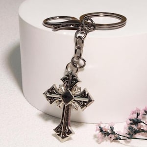 May include: A silver-toned metal keychain featuring an ornate cross pendant with intricate scrollwork detailing and a single square black stone set in the center. The cross hangs from a dark metal chain attached to a circular split ring and a lobster clasp.