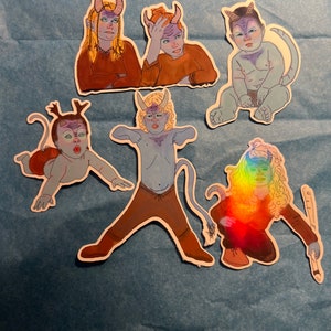 May include: Six colorful stickers featuring cartoon illustrations of blue-skinned, horned characters. The characters are in various poses, including sitting, standing, and holding a sword. The stickers are on a blue background.