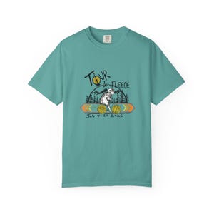 Può includere: T-shirt color verde acqua con una grafica che recita "Tour de Fleece" in corsivo nero. Il design include una pecora, un sole e il testo "July 4-26 2026". La maglietta è realizzata in un materiale morbido.