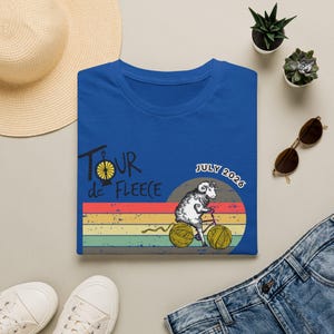 Può includere: T-shirt blu reale con una grafica di una pecora in bicicletta fatta di filato, con il testo "Tour de Fleece" e "July 2026". Il design include uno sfondo a strisce arcobaleno retrò.
