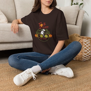 May include: A dark brown t-shirt featuring the text "Tour de Fleece" above a graphic of a sheep, yarn balls, and trees. The shirt also includes the date "July 4-24 2024". The shirt is worn with blue jeans and white sneakers.