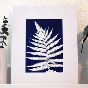 May include: Framed print with a white fern leaf design on a dark blue background. The artwork is mounted in a white mount and framed in white. Eucalyptus sprigs in a white vase are to the left of the frame.