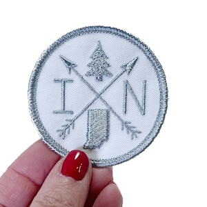 May include: A white embroidered patch with a silver outline. The patch features a stylized outline of the state of Indiana, a pine tree, and two arrows forming an 'X' with the letters 'IN' on either side of the 'X'.
