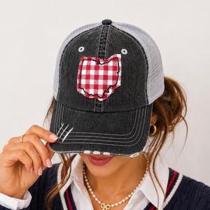 May include: A black distressed denim baseball cap with a white mesh back. The front features a red and white gingham patterned patch in the shape of the state of Ohio. The brim has frayed detailing and white stitching throughout the front panel.