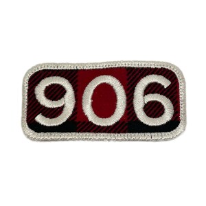 May include: Embroidered patch with the number "906" in white thread. The patch has a red and black plaid pattern background and a white border. The patch is rectangular in shape.