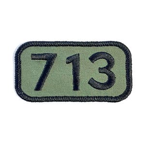 May include: An olive green patch with the number "713" embroidered in black. The patch has a black border and a rectangular shape with rounded corners. The fabric has a textured appearance.