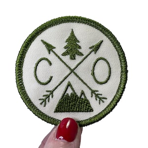 May include: Green embroidered patch with a white background. The patch features a stylized mountain, a pine tree, and two arrows that cross in the middle. The letters "CO" are on either side of the arrows.