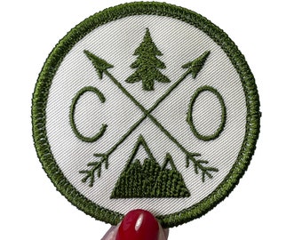 May include: A circular embroidered patch with an off-white background and a dark olive green border. The design features two crossed arrows, a pine tree, a mountain range, and the letters C and O, all stitched in dark olive green thread.