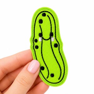 May include: A bright lime green felt patch shaped like a pickle. The patch features black embroidered outlines and scattered black embroidered dots representing bumps. Two small white embroidered highlights are positioned near the center.