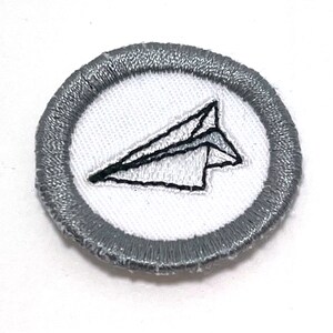 May include: A white paper airplane embroidered on a gray circle patch.