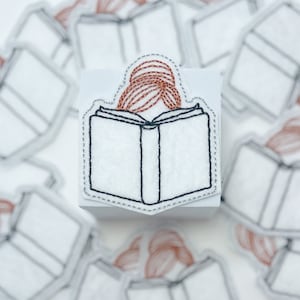 May include: A white felt patch with a black outline of a book and a person with red hair reading. The patch is surrounded by a silver stitching.