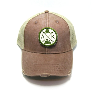 May include: Brown and beige trucker hat with a green embroidered patch that says "AK" with a mountain and arrows design.