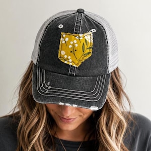 May include: A distressed black baseball cap with a gray mesh back and a frayed-edge patch on the front. The patch is mustard yellow with a white botanical floral pattern. The cap features white stitching details and a worn-in aesthetic.