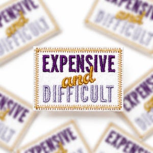 May include: A rectangular white fabric patch featuring embroidered text. The word EXPENSIVE is in purple, the word and is in gold, and the word DIFFICULT is in light purple. The patch has a gold stitched border around the entire rectangular edge.