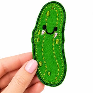 May include: A green embroidered patch shaped like a pickle with a smiling face. The design features dark green stitched outlines, light green textured details representing bumps, and black thread for the eyes and mouth with small white accents.
