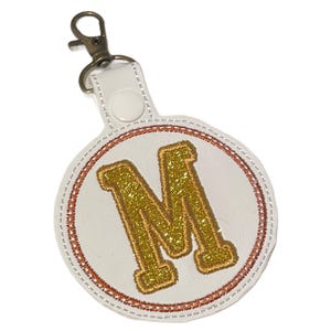 May include: White round keychain with a gold glitter letter "M" in the center. The letter is surrounded by a stitched brown border and a white background. The keychain has a silver clasp.