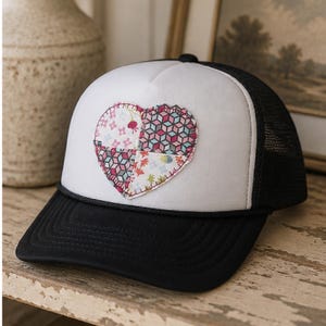 May include: A black and white trucker hat with a heart-shaped patch. The heart is made of colorful fabric pieces. The hat has a black brim and a mesh back.