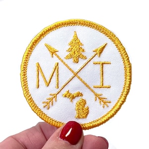 May include: Embroidered patch with a white background and a gold border. The patch features a gold tree, arrows, and the letters "MI" and "I". A gold outline of the state of Michigan is also present.