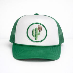 May include: A green and white trucker hat with a green mesh back and a white front panel. The front panel features a circular patch with a green cactus and a brown owl. The hat has a green brim.