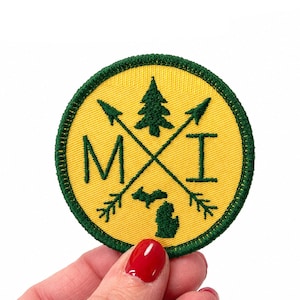 May include: A circular embroidered patch with a yellow background and a dark green border. The design features two crossed arrows, a pine tree, the letters M and I, and a silhouette of the state of Michigan in dark green embroidery thread.