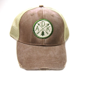 May include: A brown and beige trucker hat with a green embroidered patch that says "D.A.K." with a pine tree and crossed arrows inside a circle.