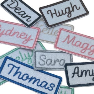 May include: A collection of rectangular white felt patches featuring embroidered names in various thread colors including black, pink, blue, silver, and green. Each patch has a matching colored border and displays names such as Dean, Hugh, Sydney, Maggie, Saro, Thomas, and Amy.