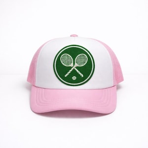 May include: A pink and white trucker hat with a green circular patch featuring crossed tennis rackets and a tennis ball. The hat has a pink brim and mesh back, and a white front panel.