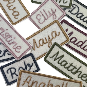May include: A collection of embroidered name patches in various colors. Each patch features a different name in a cursive font, with a contrasting border. The patches are white with gold, pink, green, and navy blue borders.