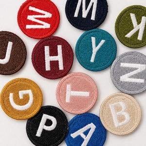 May include: A collection of circular embroidered patches in various colors, each featuring a single white capital letter. The patches include letters W, M, K, J, H, Y, N, G, T, B, P, and A, displayed in a scattered arrangement on a plain surface.
