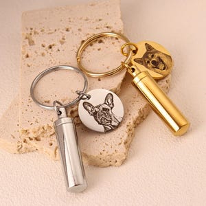 Puede incluir: Dos llaveros plateados con retratos de mascotas grabados y contenedores cilíndricos adjuntos. Un llavero presenta un Bulldog Francés, el otro un Husky.