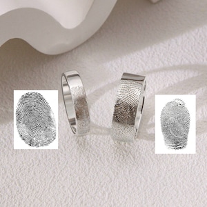 May include: Two silver rings featuring fingerprint patterns. One is a plain band, the other has a flat top. Two black and white fingerprint images are also shown. The rings are presented on a white surface.