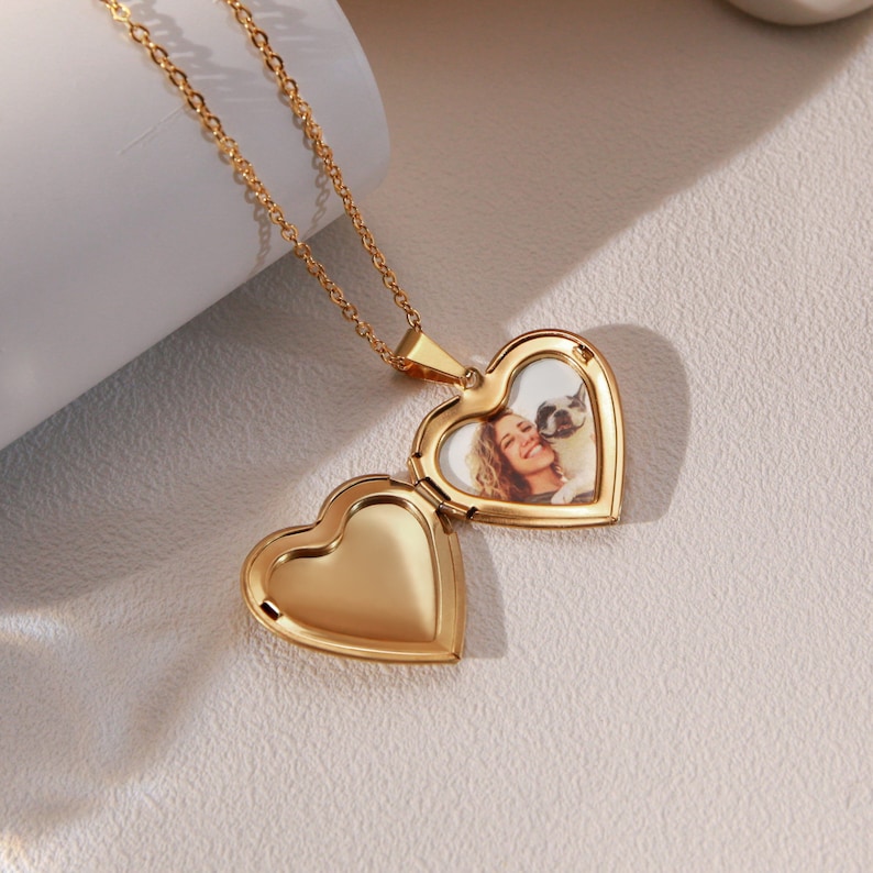 May include: Gold heart-shaped locket necklace with a photo of a woman and a dog inside. The locket is open, revealing the photo.