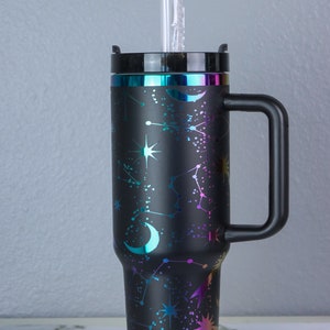 May include: A black stainless steel travel mug with a constellation and moon design. The mug has a handle and a lid. A clear plastic straw is in the mug.