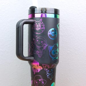以下が含まれることがあります： ハンドルと蓋が付いた黒いトラベルマグ。マグカップは、ホログラフィックなポケモンのキャラクターとモンスターボールのパターンで覆われています。